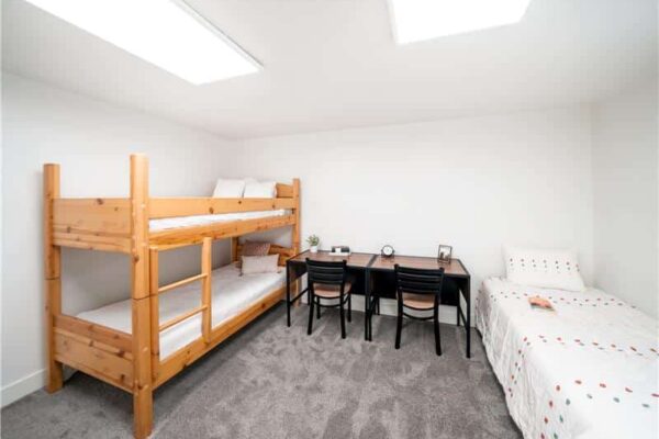 Bright dorm-style room with a wooden bunk bed on the left, a desk and four chairs in the center, and a single bed on the right under skylight windows.